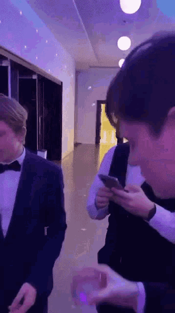 a man in a tuxedo is looking at a cell phone while another man looks on .