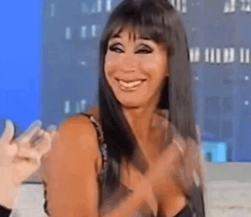 a woman with long black hair and a plunging neckline is smiling and clapping her hands .