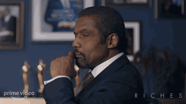 a man in a suit and tie is sitting in front of a wall with trophies and the word riches on it
