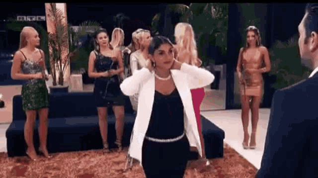 a woman in a white jacket is dancing in front of a group of women in dresses .