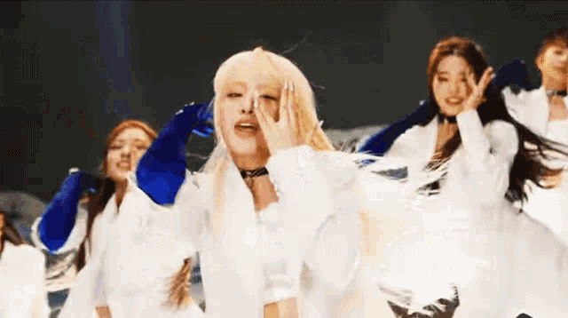 a group of women are dancing together on a stage while wearing white dresses and blue gloves .