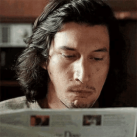 a man with long hair is reading a newspaper in a kitchen .