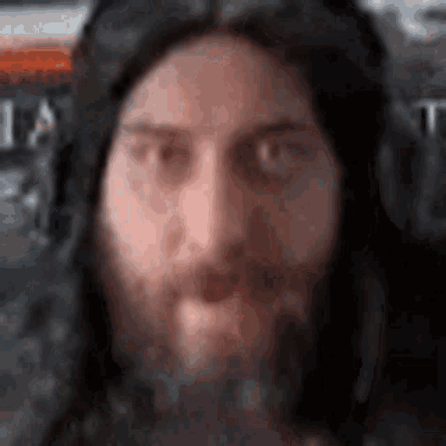 a man with long hair and a beard is looking at the camera with a blurry background .