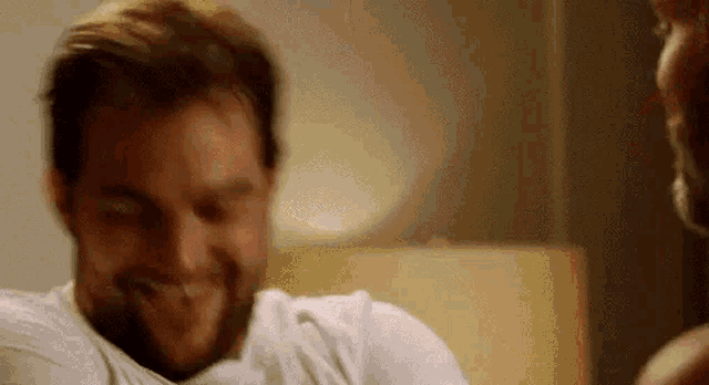 a man with a beard is laughing while sitting on a bed next to another man .