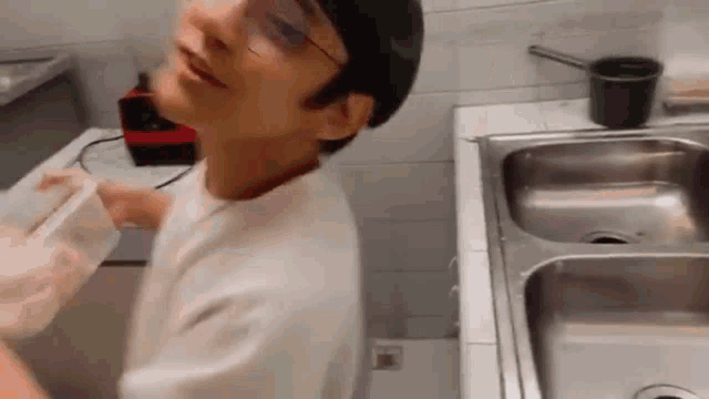 a man wearing glasses is standing in front of a kitchen sink holding a plastic container .