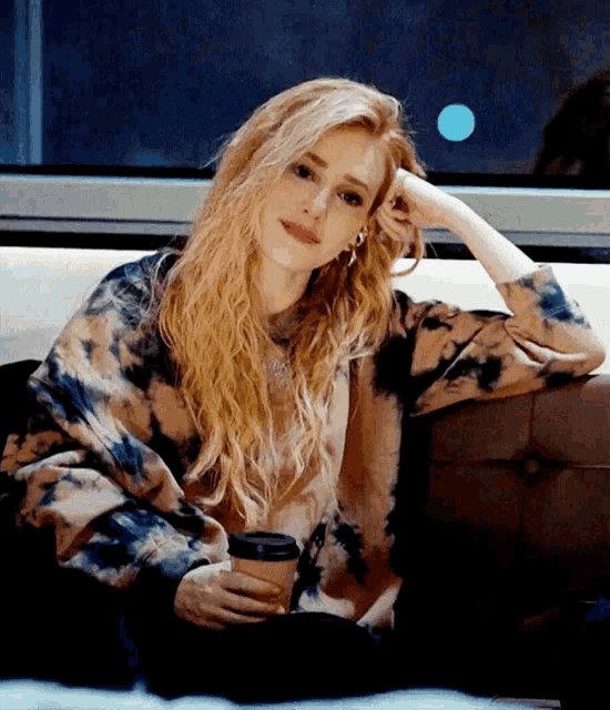 a woman in a tie dye sweater is sitting on a couch holding a cup of coffee