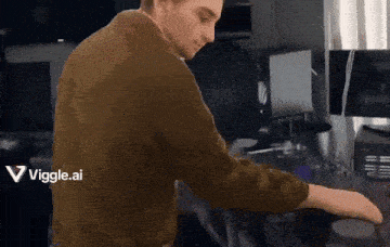 a man in a brown sweater is standing in front of a desk with a viggle.ai logo on the bottom