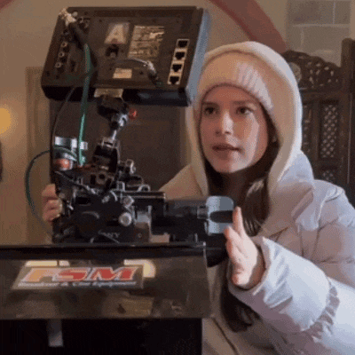 a woman in a hooded jacket is holding a camera in front of a machine that says fsm