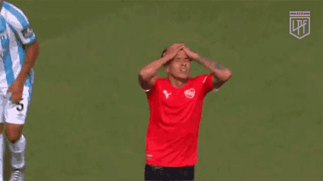 a soccer player in a red shirt holds his head in his hands while another player in a blue shirt looks on .