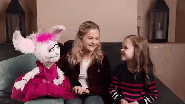 two little girls are sitting on a couch with a puppet in a pink dress .