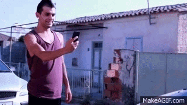 a man in a purple tank top is holding a cell phone in front of a white car .