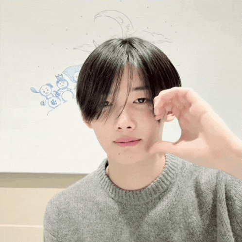 a young man wearing a grey sweater is making a heart shape with his hands