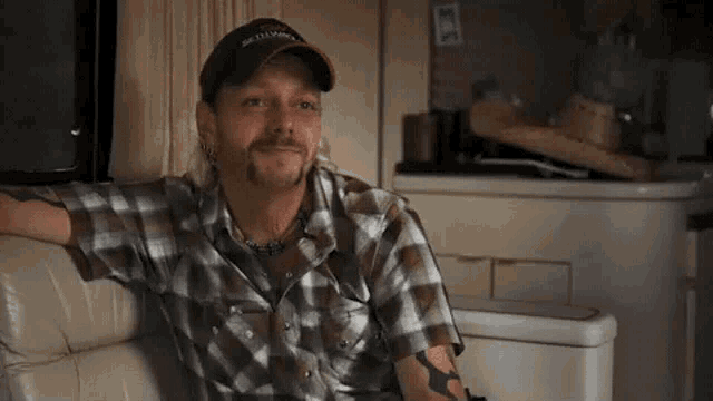 a man with a beard is sitting on a couch wearing a plaid shirt and a hat .