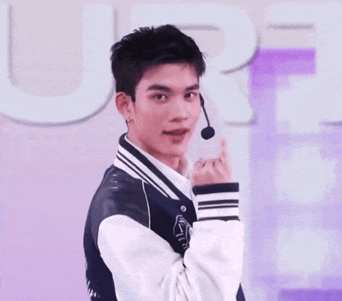 a young man wearing a bomber jacket and a microphone is making a heart shape with his hands .