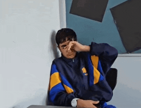 a young man in a blue and yellow jacket is sitting at a desk covering his face with his hand .