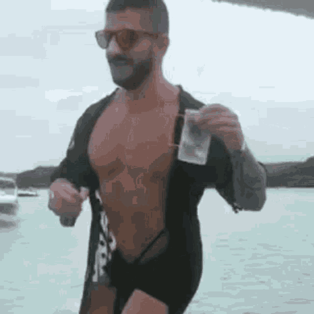 a shirtless man is walking on a boat in the water holding a drink .