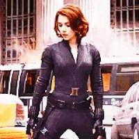 a woman in a black suit is standing on a city street with a gun .