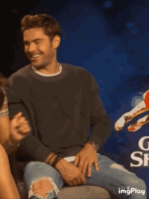 a man in a black sweater and ripped jeans is smiling while sitting in front of a sign that says imgplay
