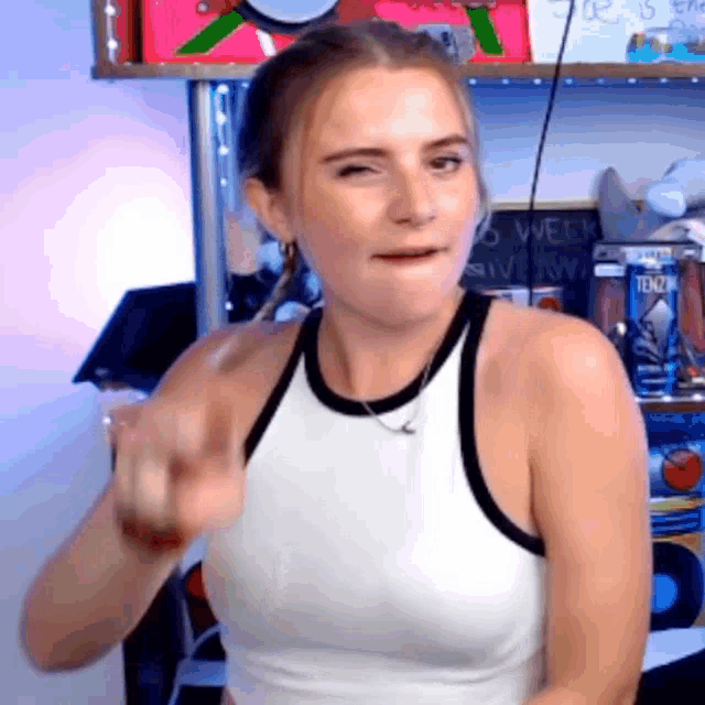 a woman in a white tank top is making a funny face while pointing at the camera .