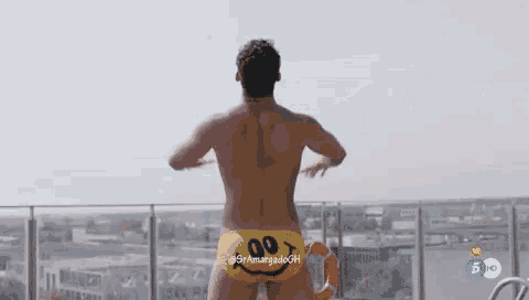 a shirtless man wearing a yellow swim trunk with a smiley face on it is standing on a balcony .