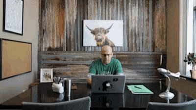 a man sitting at a desk with a laptop and a picture of a cow on the wall