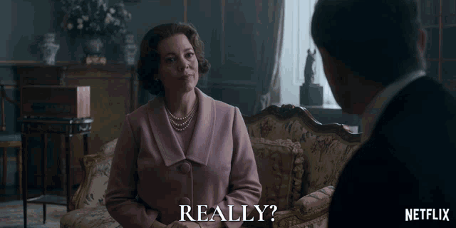 a woman in a pink coat is sitting on a couch talking to a man in a suit and says really