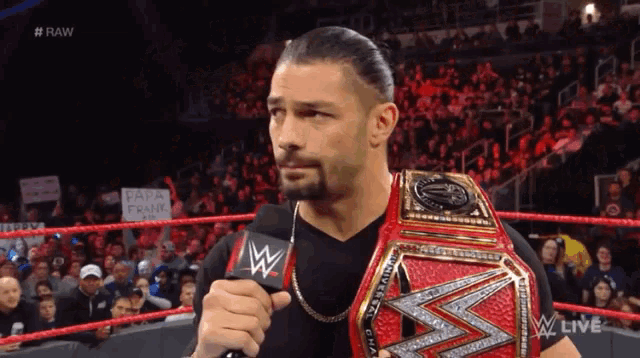 roman reigns is holding a microphone in his hand while wearing a wrestling belt .