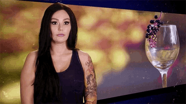 a woman with a tattoo on her arm is standing in front of a wine glass with grapes on it .