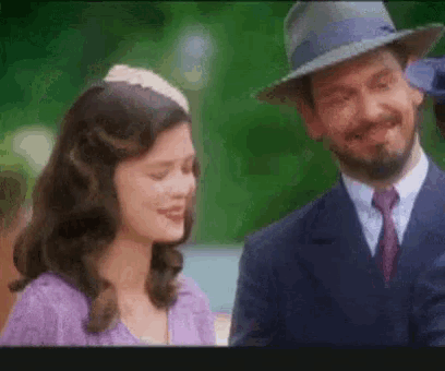 a man in a suit and tie is standing next to a woman in a purple dress and hat .
