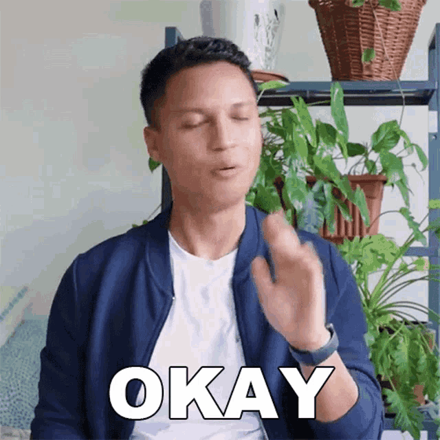 a man in a blue jacket says okay in front of a plant shelf