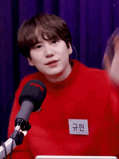 a young man wearing a red sweater stands in front of a microphone with the word now on it
