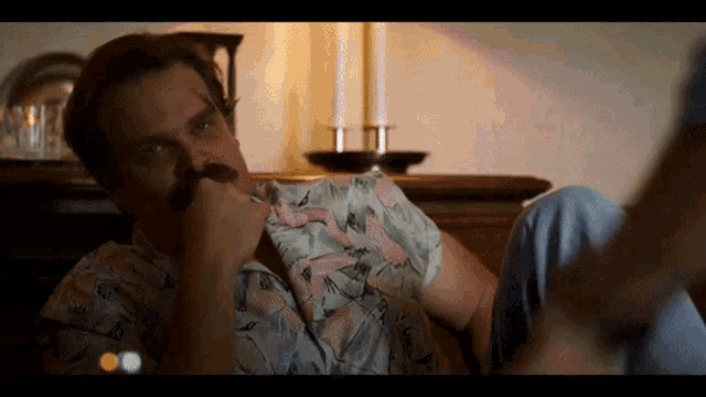 a man wearing a flamingo shirt is sitting on the floor with his hand on his face .