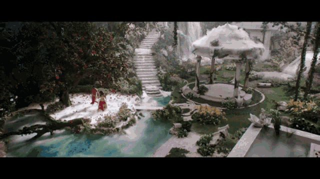 a woman in a red dress stands in front of a waterfall in a garden