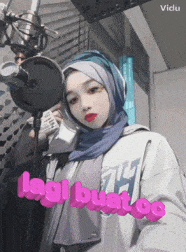 a woman wearing a hijab stands in front of a microphone with the words lagi buat oe written in pink