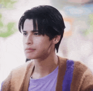 a young man wearing a purple t-shirt and a brown cardigan is looking at the camera .