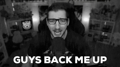a man wearing glasses is talking into a microphone and the words `` guys back me up '' are visible .