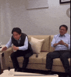 two men are sitting on a couch and one is holding a remote control