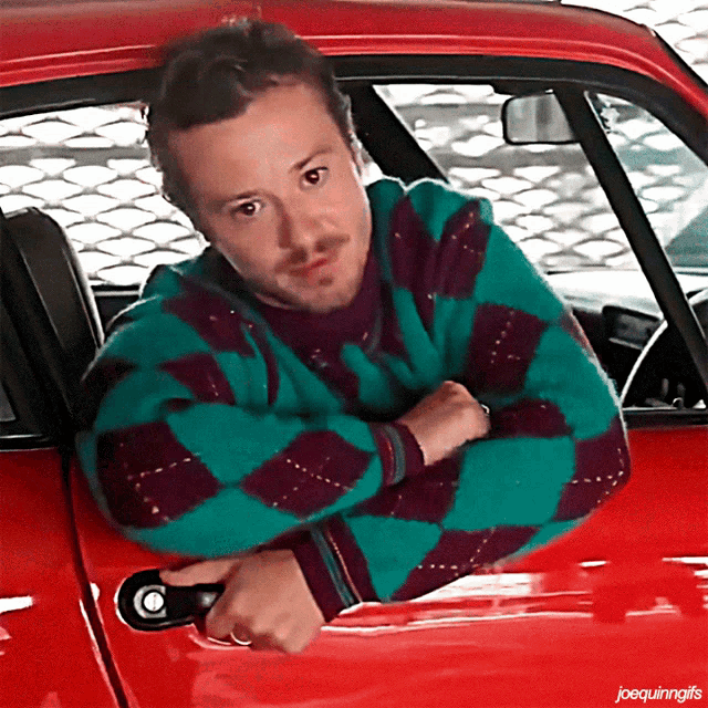 a man in a green and purple plaid sweater leans out of a red car