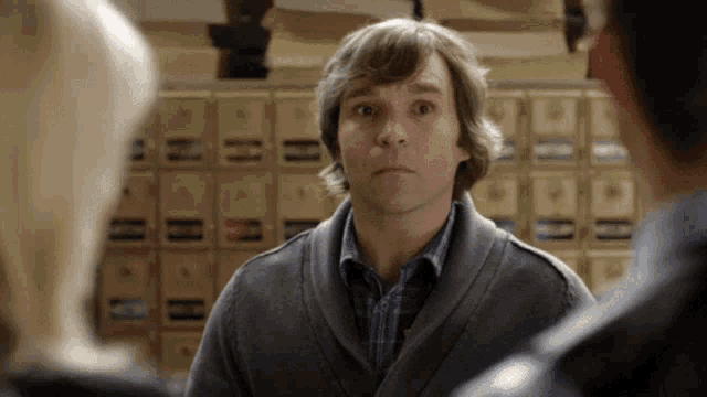 a man in a grey sweater stands in front of a wall of mailboxes with the letter h on them