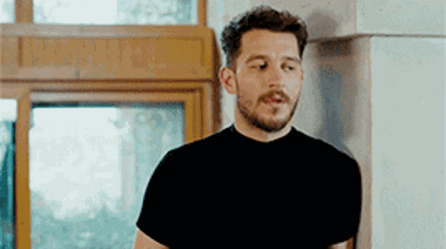 a man with a beard and mustache is wearing a black shirt and standing in front of a window .