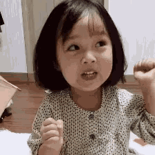 a little girl in a polka dot shirt is sitting on the floor with her fist in the air .