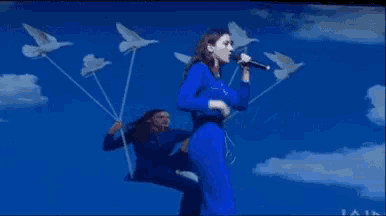 a woman in a blue jumpsuit sings into a microphone while another woman sits on a swing
