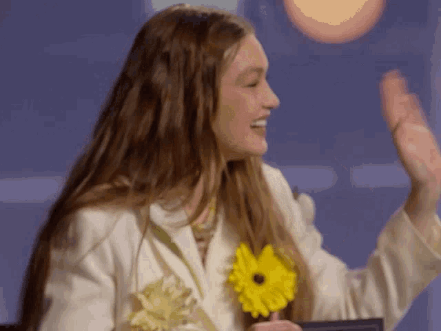 a woman in a white jacket is holding a yellow flower in her hand and smiling .