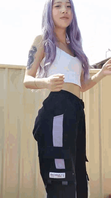 a woman with purple hair is wearing a white crop top and black pants
