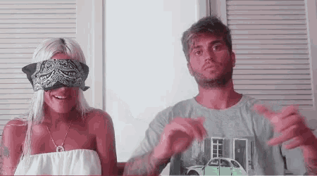 a man and a woman are standing next to each other . the woman is wearing a bandana on her eyes .