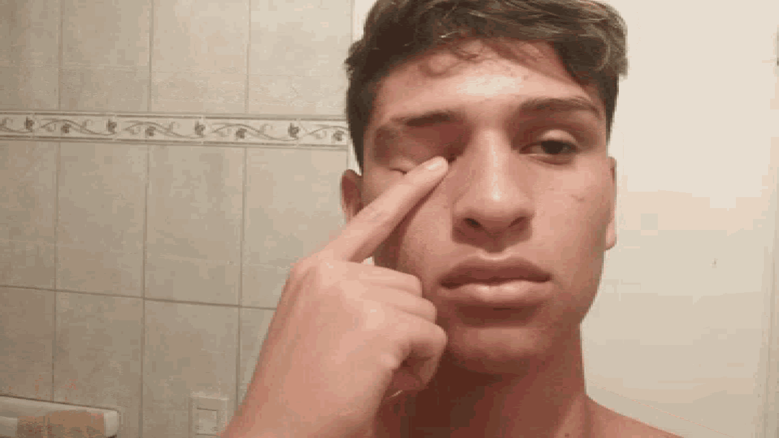 a young man is pointing at his nose with a tiled wall in the background