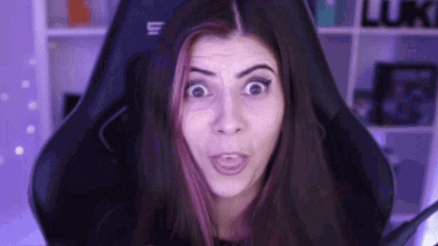 a woman with purple hair is sitting in a gaming chair and making a surprised face .