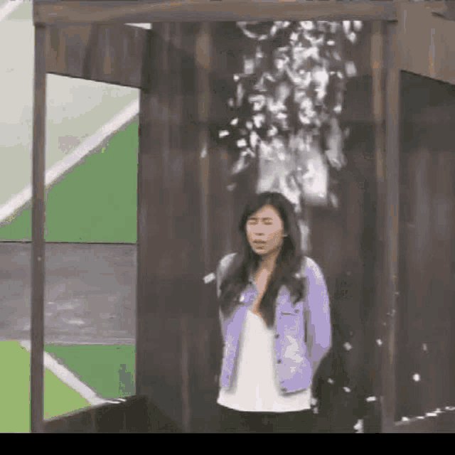 a woman in a purple jacket is standing in front of a wooden wall with confetti falling from it .