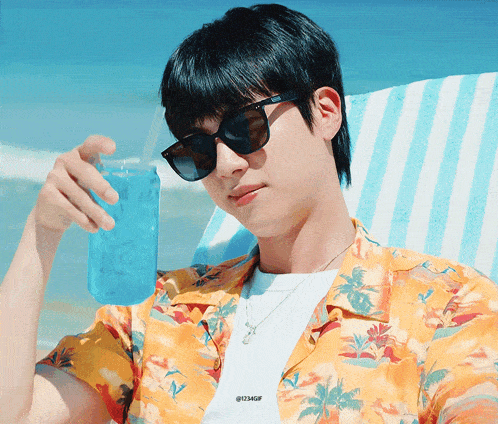 a man wearing sunglasses sits on a beach holding a can of blue liquid