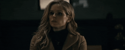 a woman in a brown coat and black turtleneck is looking at the camera